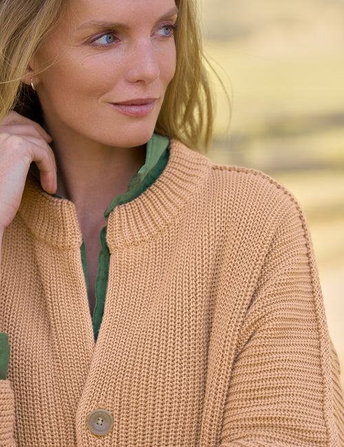 A close-up of a woman wearing a chunky knit cardigan in a warm camel tan color, layered over a green collared shirt. The background is a soft, blurred outdoor setting in warm beige tones., view 3