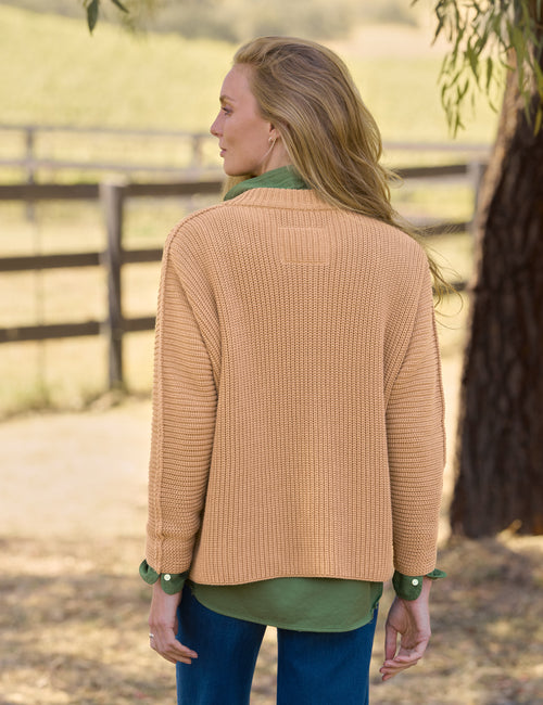 A woman shown from the back wearing a ribbed knit cardigan in a warm camel tan color over an olive green shirt, paired with blue jeans, set against a blurred outdoor background with a fence and tree., alternate view