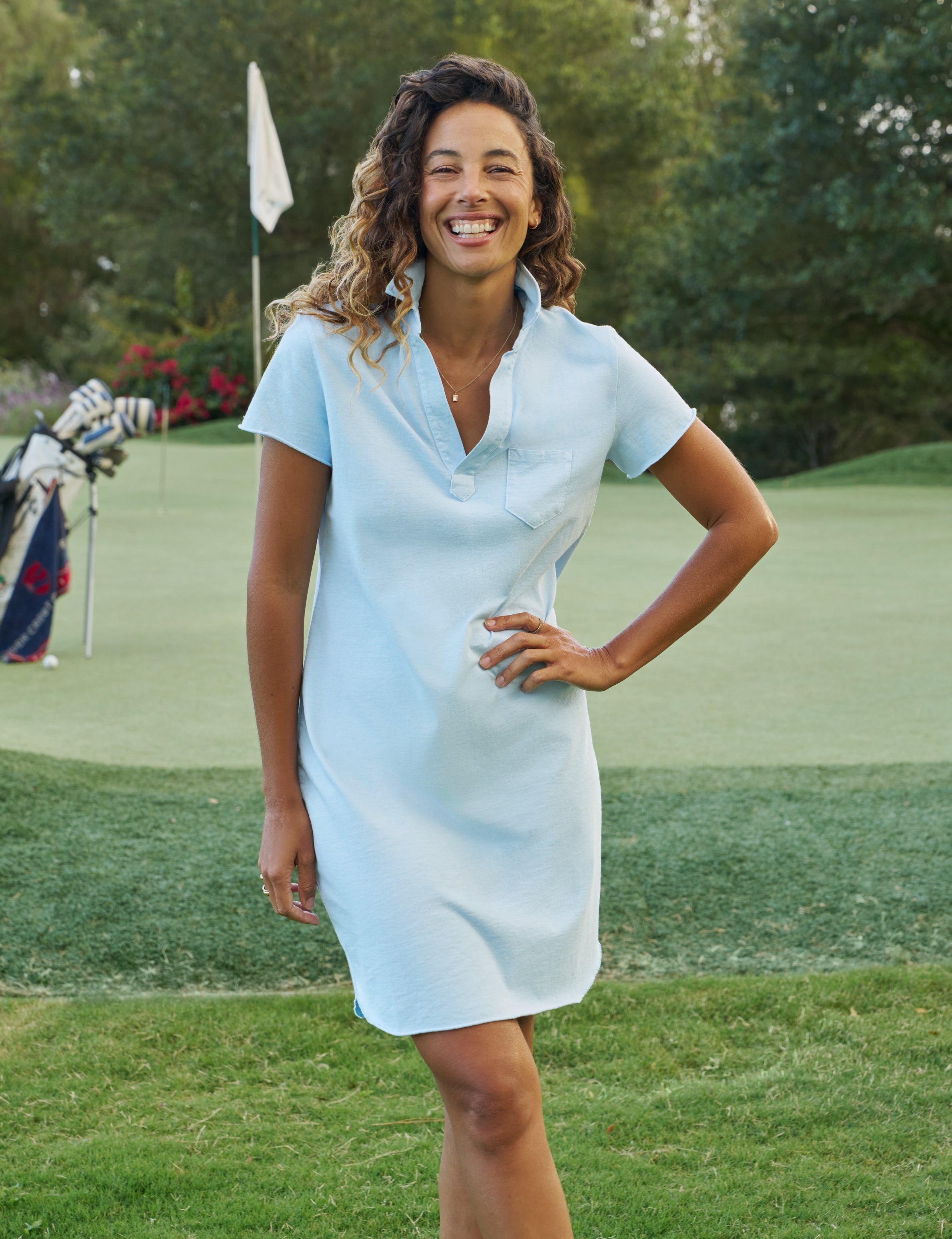 Woman in a light blue dress standing on a golf course with a golf club and flag in the background.