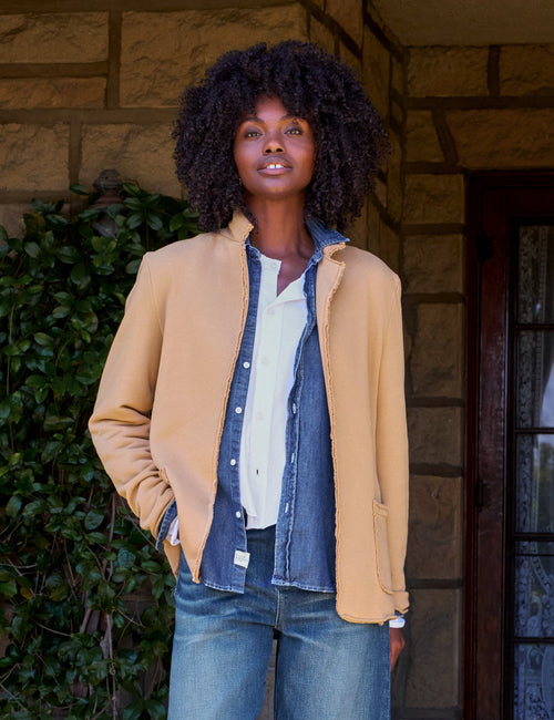 A woman stands on a stone porch wearing a tan blazer over a white shirt and denim shirt, paired with blue jeans and dark loafers., front view