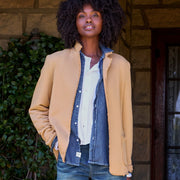 A woman stands on a stone porch wearing a tan blazer over a white shirt and denim shirt, paired with blue jeans and dark loafers.