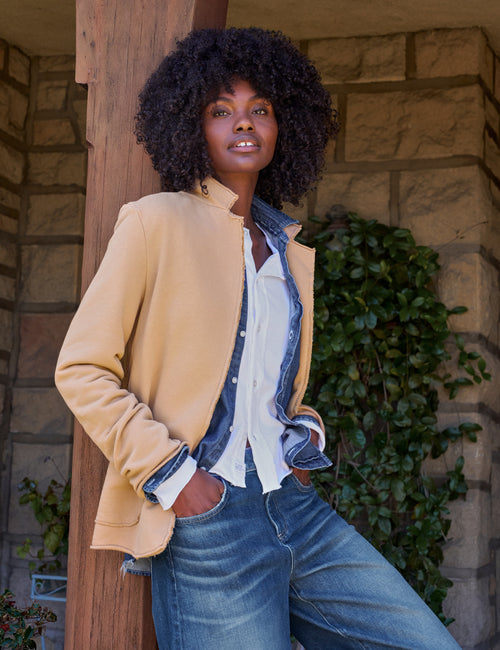 A woman stands outdoors against a stone wall and wooden post, wearing a tan jacket over a white button-up shirt and blue jeans., alternate view