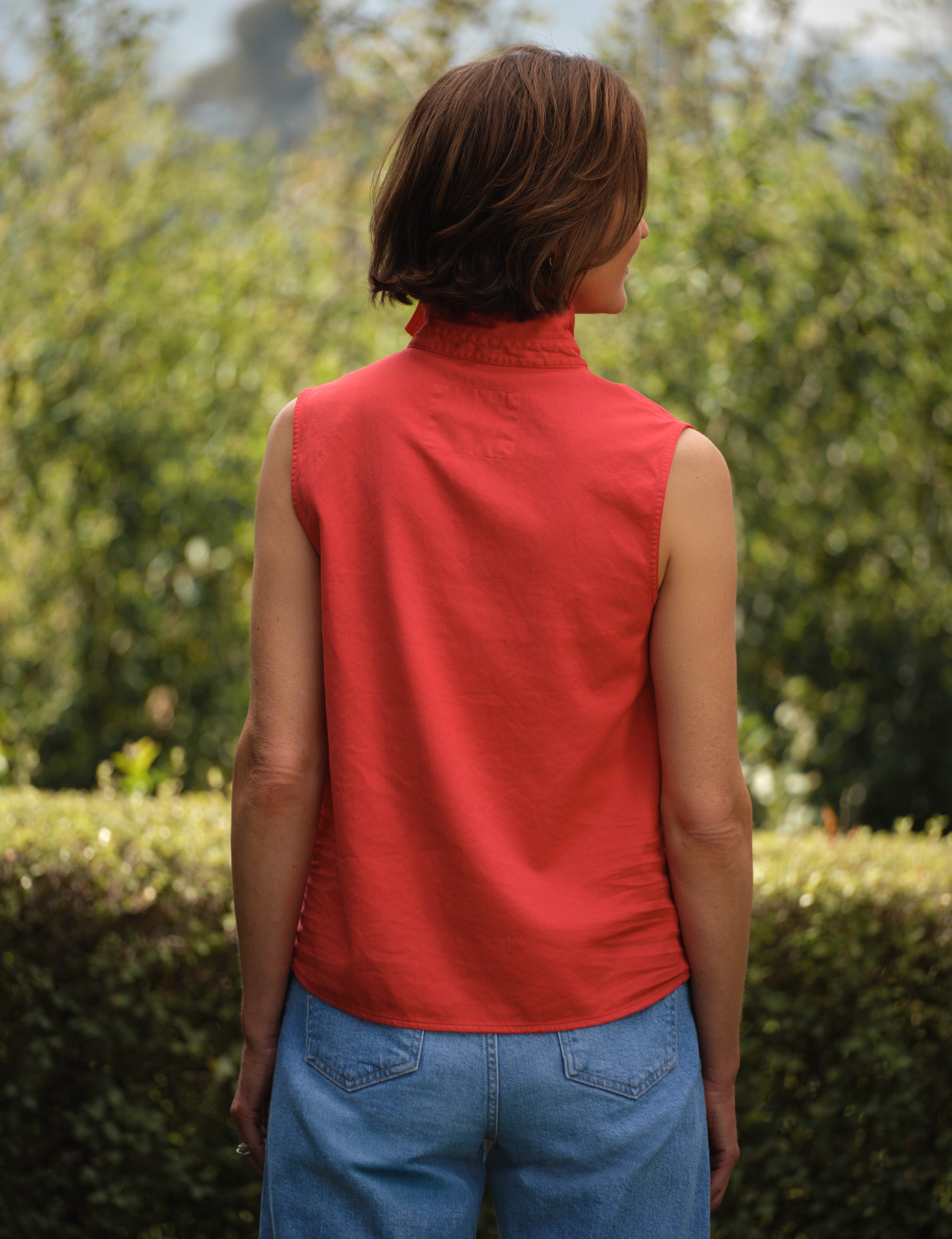 Person wearing a red sleeveless top and blue jeans standing outdoors with greenery in the background, view 4