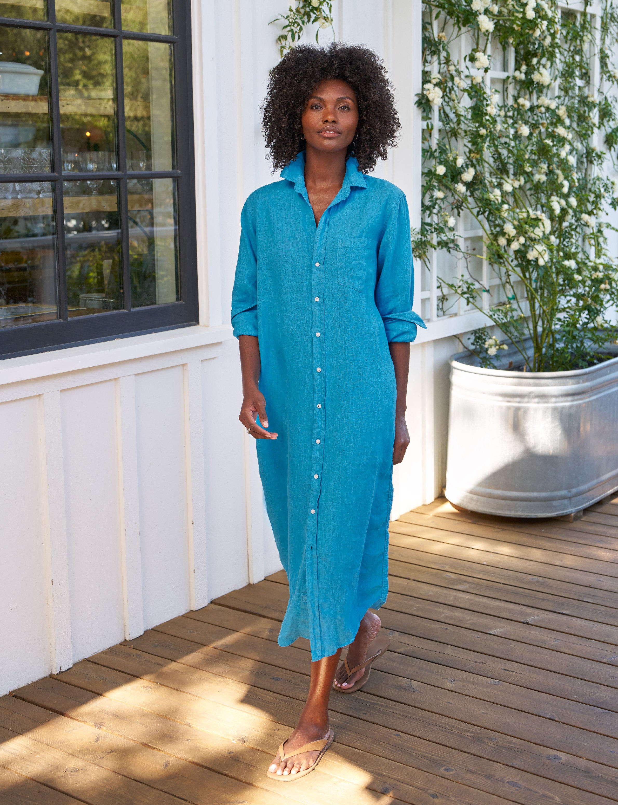 Woman wearing a blue dress standing on a wooden deck with plants in the background, front view