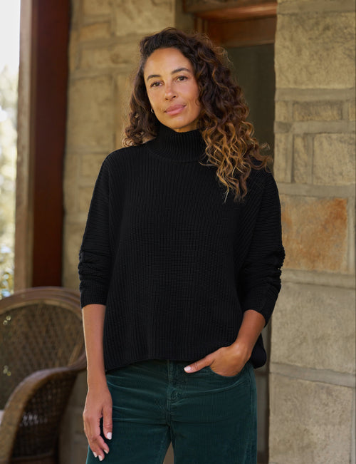 A woman with curly brown hair wearing a black ribbed turtleneck sweater and dark green pants, standing in front of a stone wall., front view