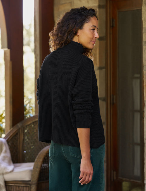 A woman wearing a black ribbed turtleneck sweater and teal jeans, standing on a stone porch with a wicker chair in the background., alternate view