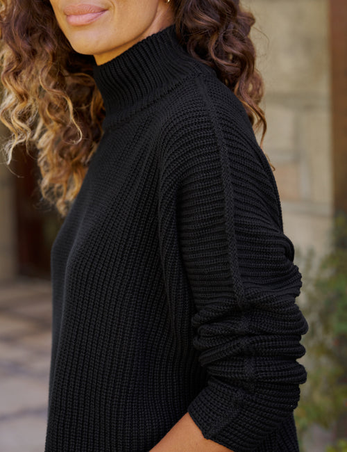A close-up side view of a woman wearing a black ribbed knit mock neck sweater, showing the texture of the fabric and her curly brown hair against a blurred outdoor background., view 3