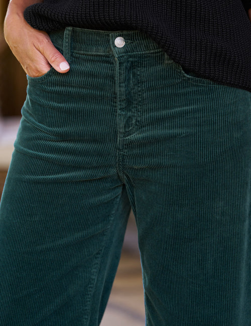 Close-up of a person wearing dark green corduroy pants with a black ribbed sweater, showing the waistband, button, and front pocket area., view 3