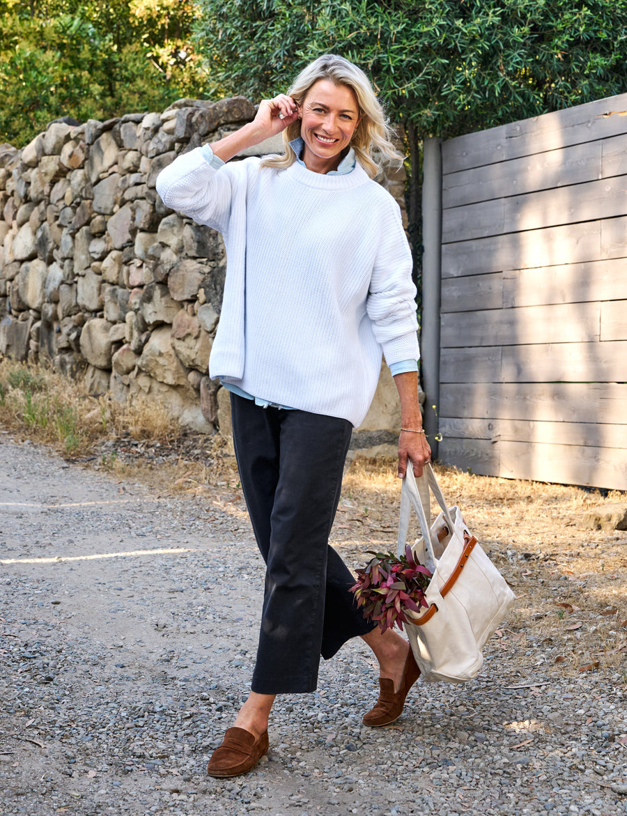 Woman in a white sweater and dark pants walking outdoors with a bag.