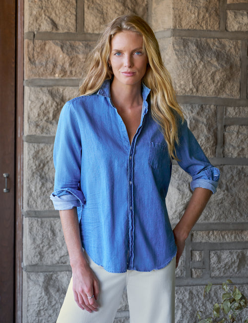A woman with long blonde hair wearing a blue chambray button-down shirt and white pants, standing in front of a rustic stone wall., front view