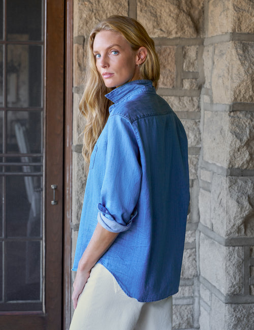 A woman with blonde hair stands in profile wearing a blue denim shirt with rolled-up sleeves and off-white pants, positioned in front of a stone wall and a dark wooden door., alternate view