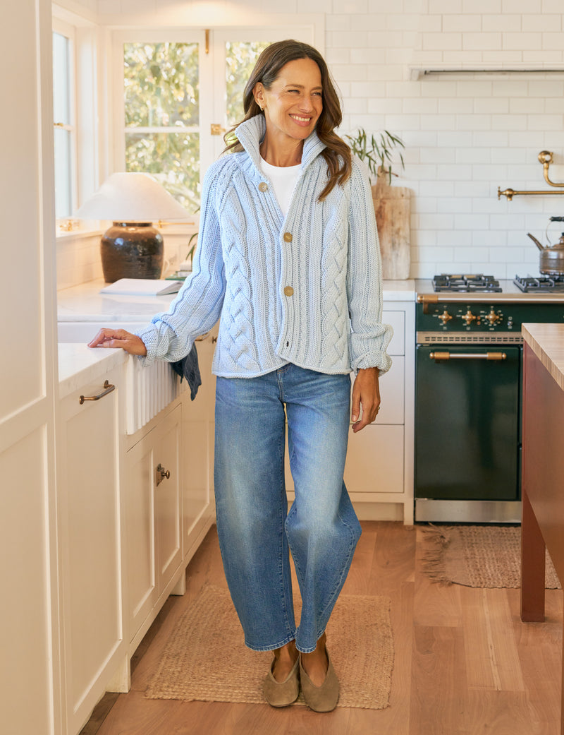 Woman standing in a kitchen wearing a light blue cardigan and jeans.