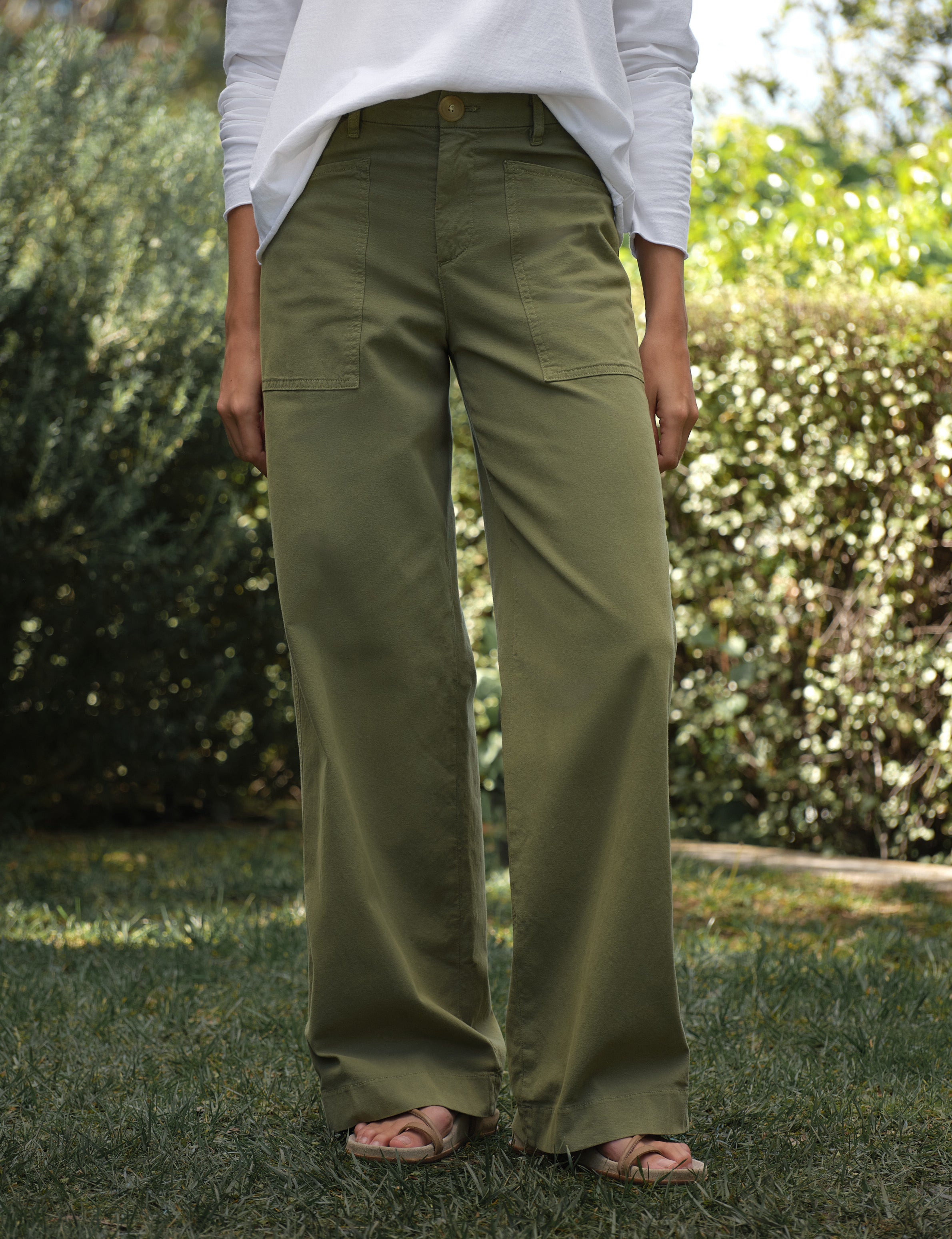 Person wearing olive green pants and a white shirt standing outdoors with greenery in the background, front view