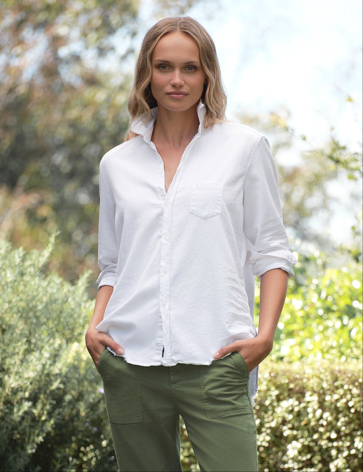Woman wearing a white shirt and green pants standing outdoors with greenery in the background, front view