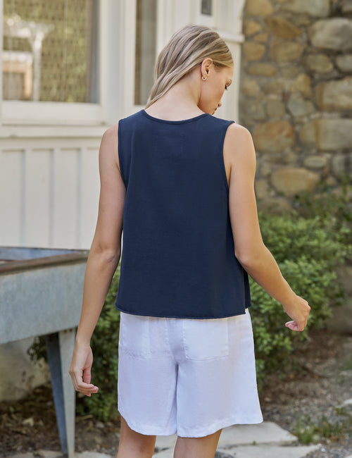 Woman wearing a navy sleeveless top and white shorts standing outdoors near a stone wall., alternate view