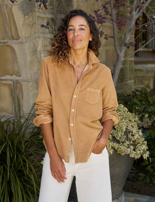 A woman wearing a tan corduroy button-down shirt with white pants, standing outdoors in front of a stone wall and plants., front view