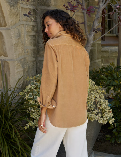 A woman wearing a tan corduroy button-up shirt and white pants, photographed outdoors in front of a stone wall and plants., alternate view