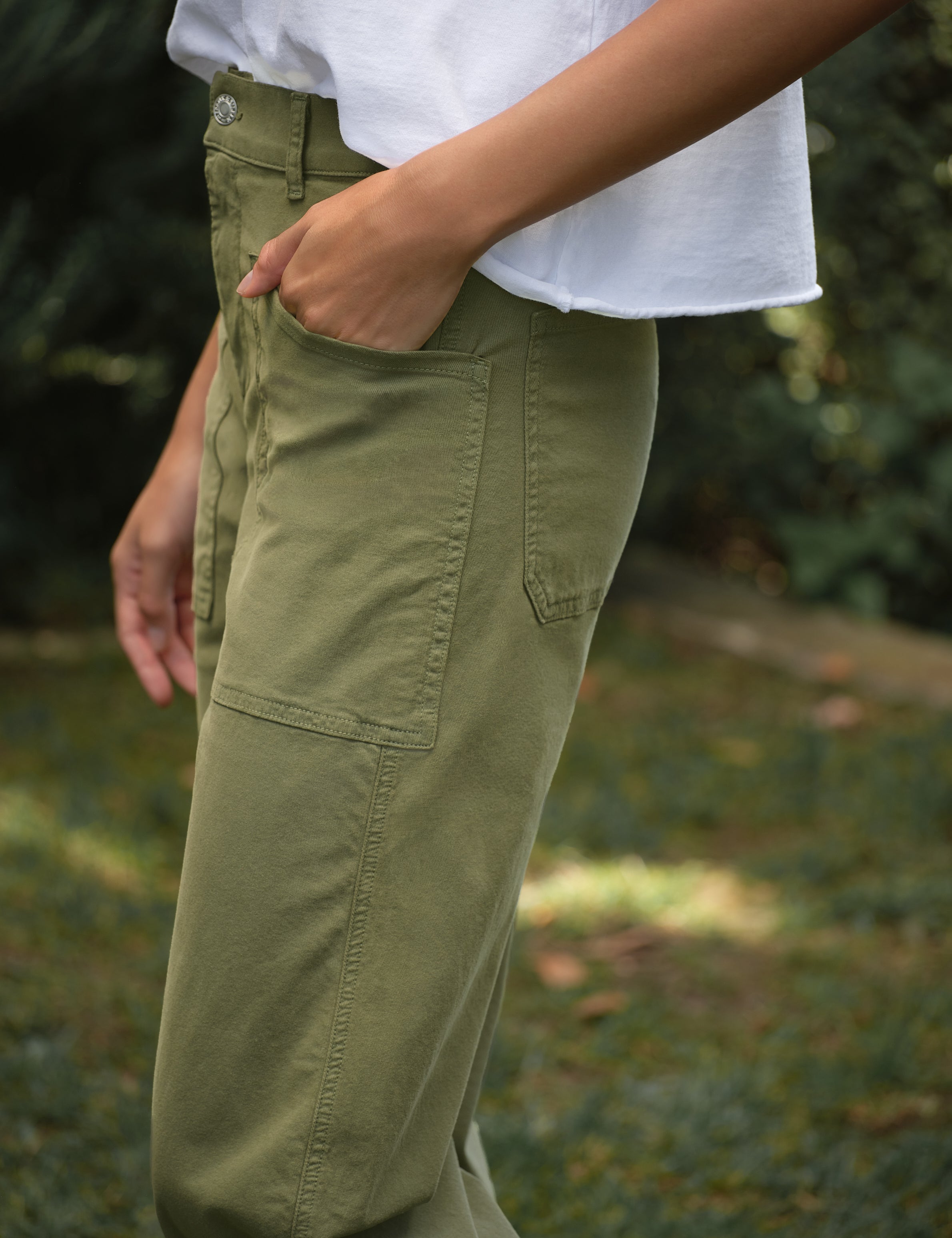 Person wearing green pants and a white shirt with a blurred natural background, view 3