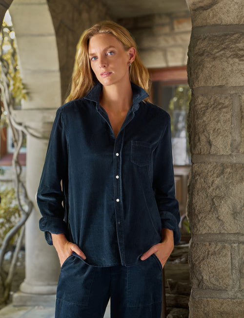 A woman wearing a dark navy corduroy button-up shirt and matching corduroy pants, standing outdoors in front of a stone archway., front view