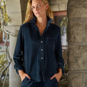A woman wearing a dark navy corduroy button-up shirt and matching corduroy pants, standing outdoors in front of a stone archway.