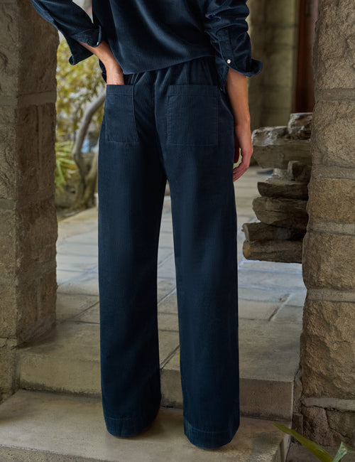 A person wearing dark navy wide-leg trousers and a matching dark top, standing in a stone doorway or porch setting., view 4