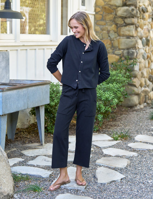 Woman in navy outfit standing outdoors near a stone wall and garden., front view