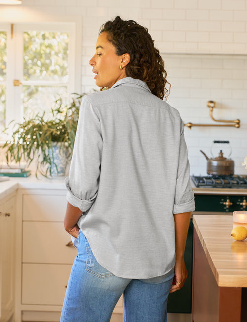 Woman standing in a kitchen wearing a light gray shirt and blue jeans., alternate view