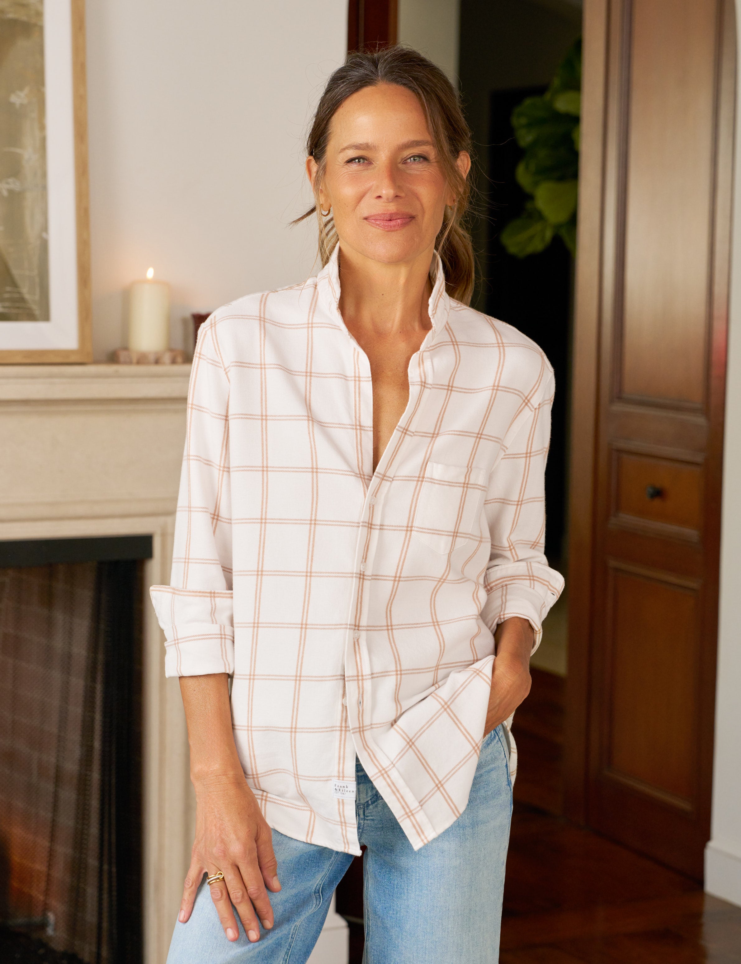 Woman wearing a white plaid shirt and blue jeans standing in a room with a fireplace.