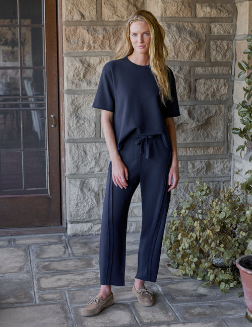 A woman stands on a stone patio wearing a dark navy short-sleeve top and matching navy barrel-leg pants with a drawstring waist, paired with tan lace-up shoes., front view