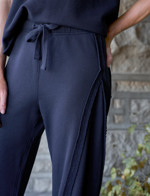 Close-up of a person wearing dark navy wide-leg sweatpants with a drawstring waist and side seam detailing, standing outdoors against a stone wall., view 3