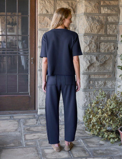 A woman stands on a stone porch wearing a dark navy blue short-sleeve top and matching barrel-leg trousers, viewed from the back., alternate view