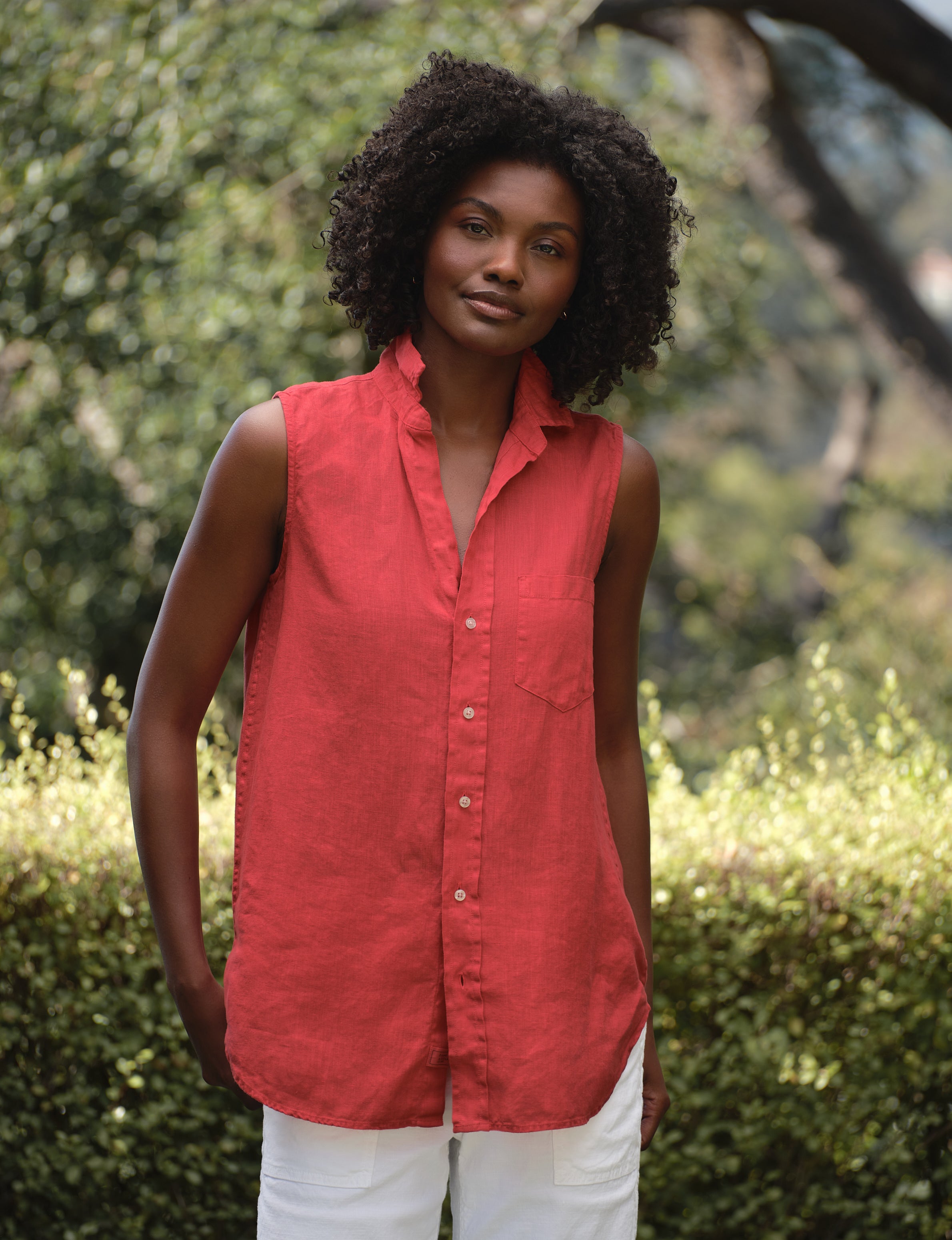 Woman wearing a red sleeveless shirt standing outdoors with greenery in the background, front view