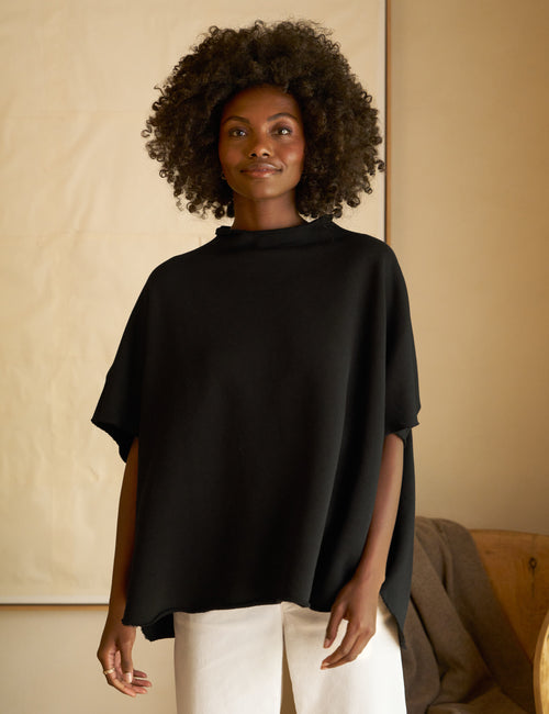 A woman wearing a black funnel neck capelet with short sleeves over a white top, paired with white trousers., front view