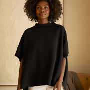 A woman wearing a black funnel neck capelet with short sleeves over a white top, paired with white trousers.