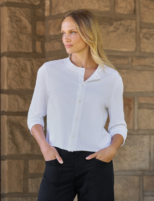 A woman wearing a white long-sleeve button-up shirt and black pants, standing against a rustic stone wall., front view