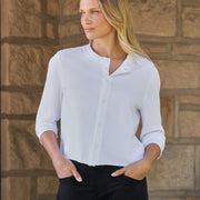 A woman wearing a white long-sleeve button-up shirt and black pants, standing against a rustic stone wall.
