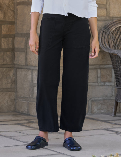 A person wearing black barrel-leg cropped pants with a white long-sleeve top and dark loafers, standing outdoors against a stone wall., front view