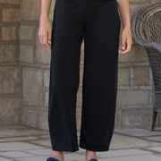 A person wearing black barrel-leg cropped pants with a white long-sleeve top and dark loafers, standing outdoors against a stone wall.