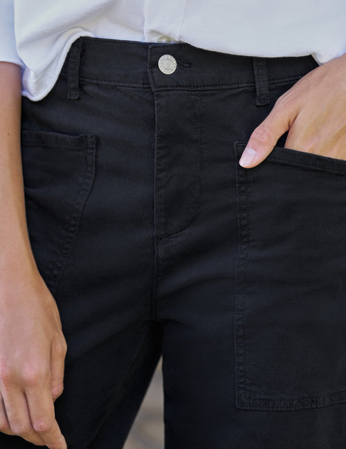 Close-up of a person wearing black pants, showing the front waistband with a silver button, side pockets, and a hand tucked into the right pocket., view 3