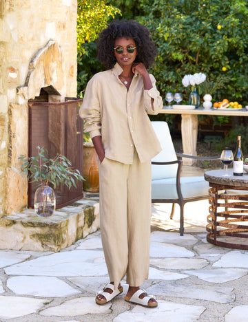 A woman standing in an outdoor setting wearing a beige 'Eileen button-up' and 'Maisie wide-leg pant' from the TUSCANY GETAWAY SET.