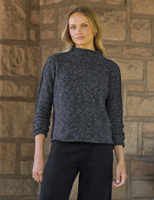 A woman wearing a dark charcoal gray marled knit mock neck sweater and black pants, standing in front of a rustic stone wall., front view