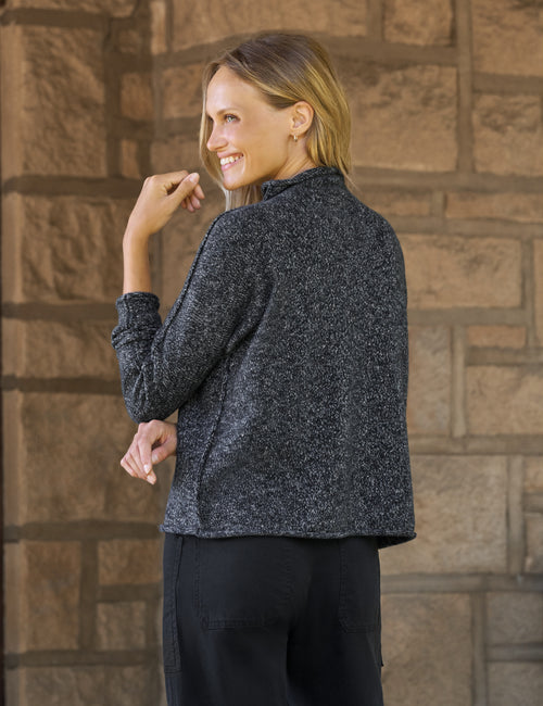 A woman wearing a dark gray marled knit long-sleeve cropped sweater, shown from the back, standing against a brown stone wall., alternate view