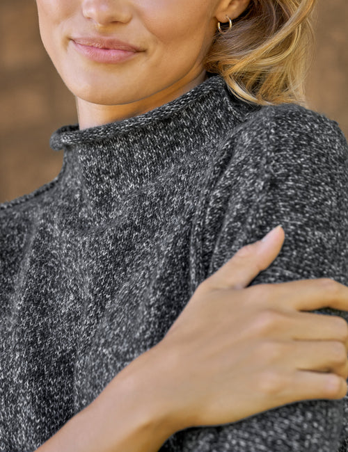 Close-up of a woman wearing a dark gray and white heathered knit mock neck sweater, with her hand resting on her shoulder., view 3