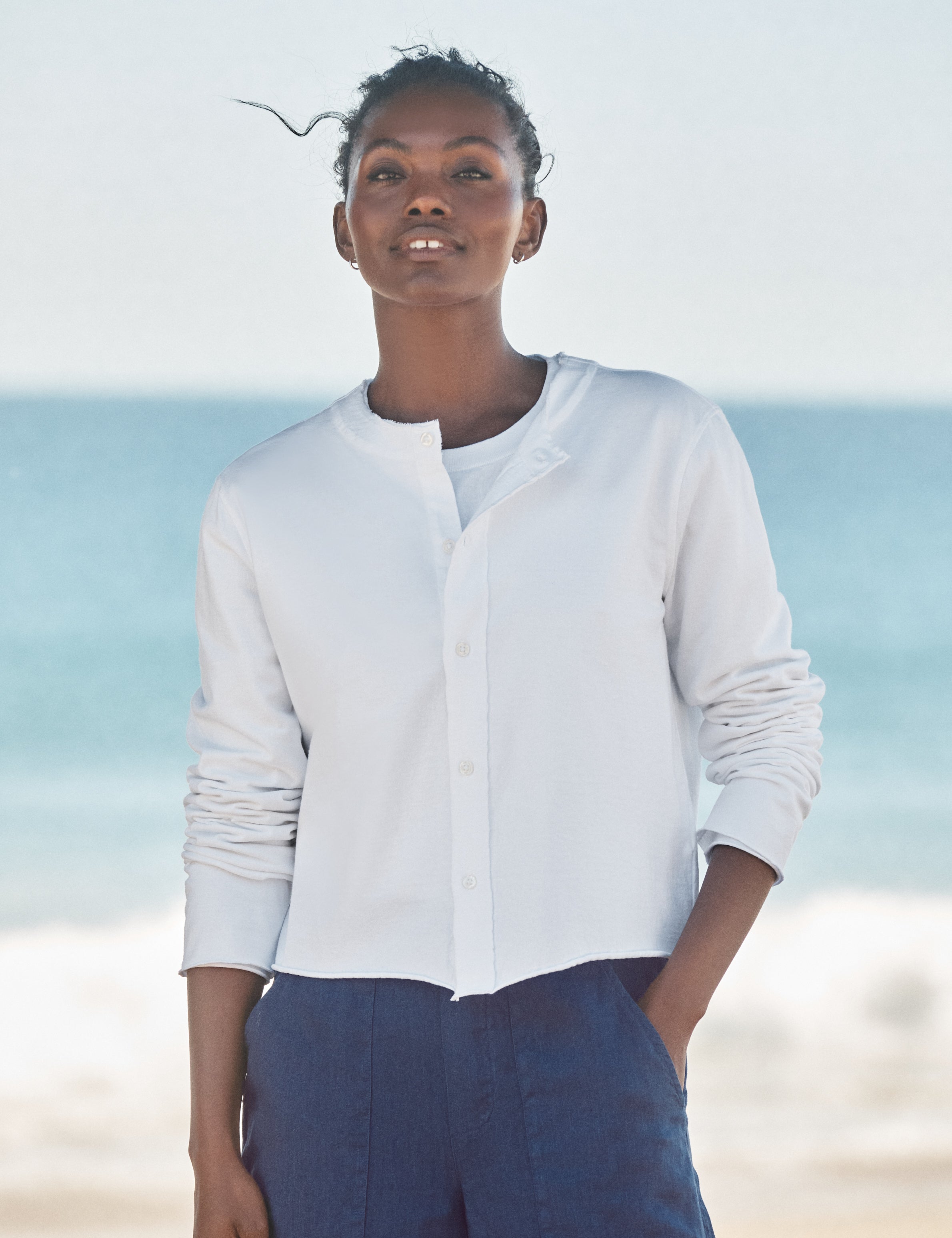 Woman wearing a light-colored cardigan and dark pants standing on a beach with ocean in the background, view 4