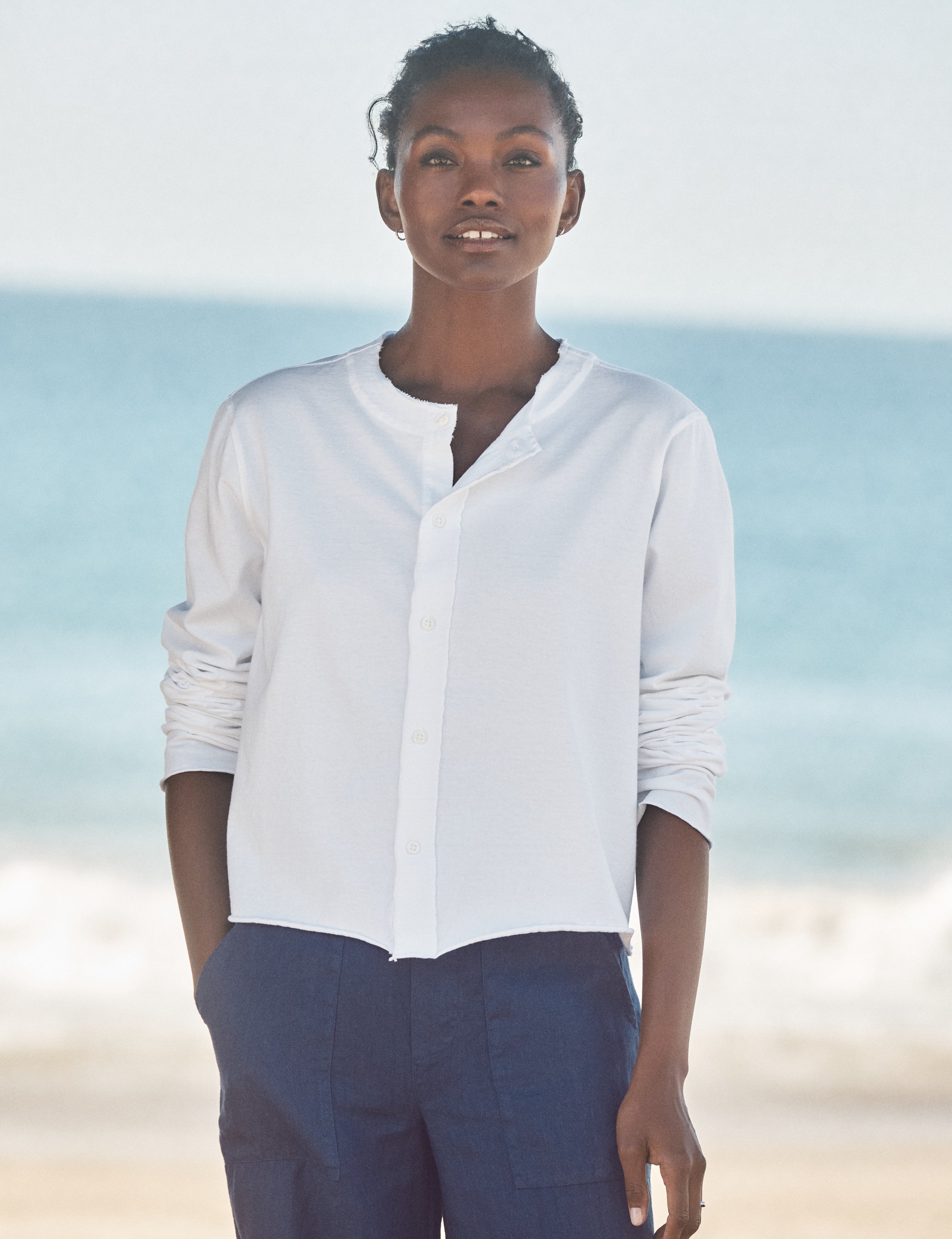 Person wearing a white shirt and dark pants standing on a beach with ocean in the background, front view