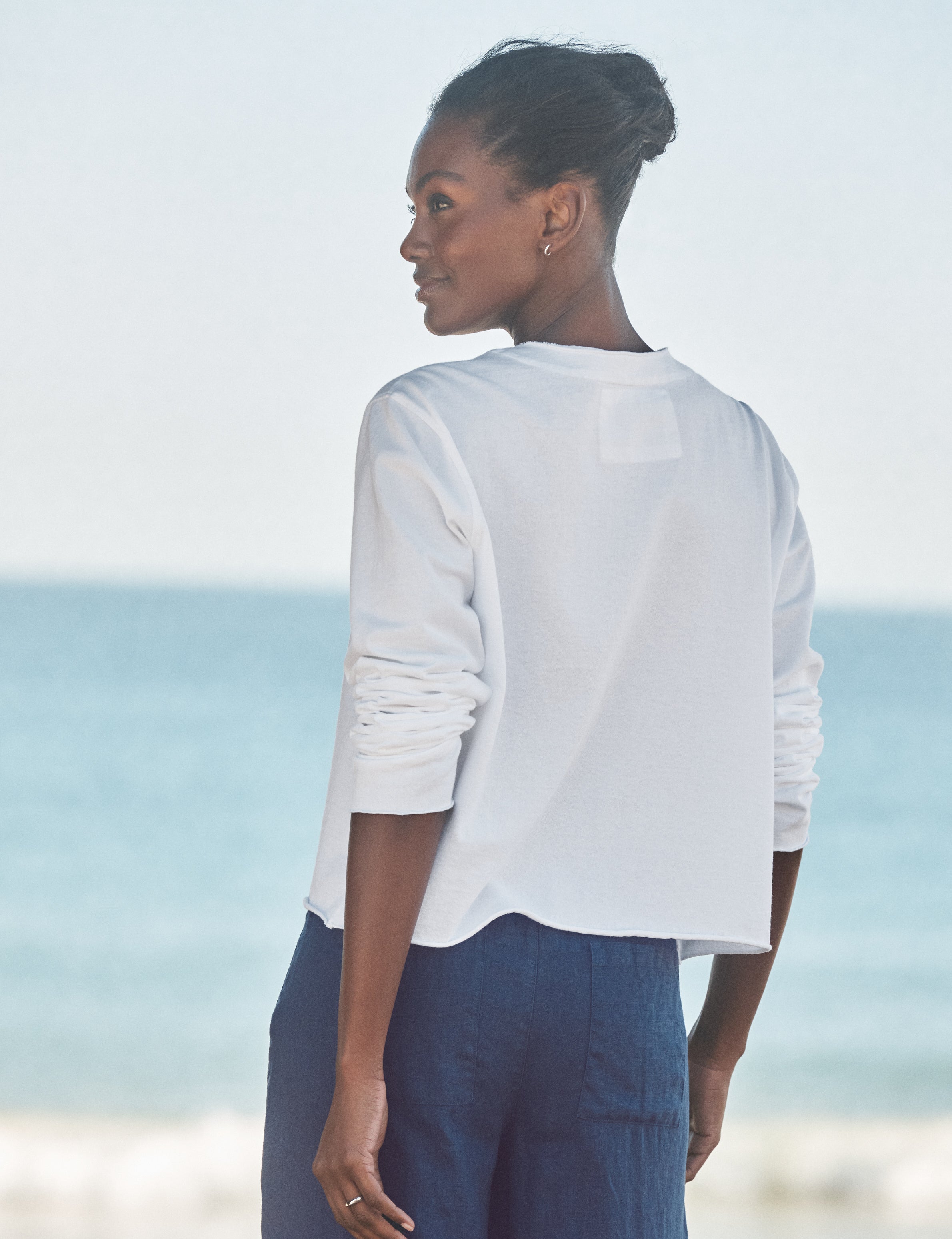Woman wearing a white top and blue pants standing on a beach., alternate view