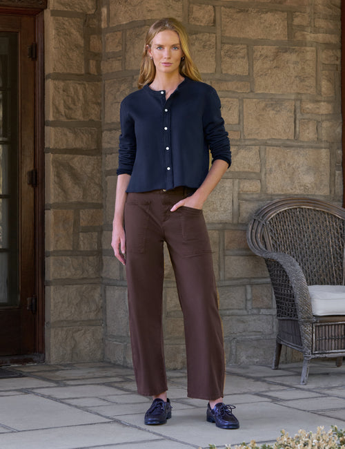 A woman stands on a stone patio wearing a navy blue long-sleeve button-front top, brown barrel-leg cropped pants, and navy lace-up shoes. A wicker chair is visible against a stone wall background., view 5