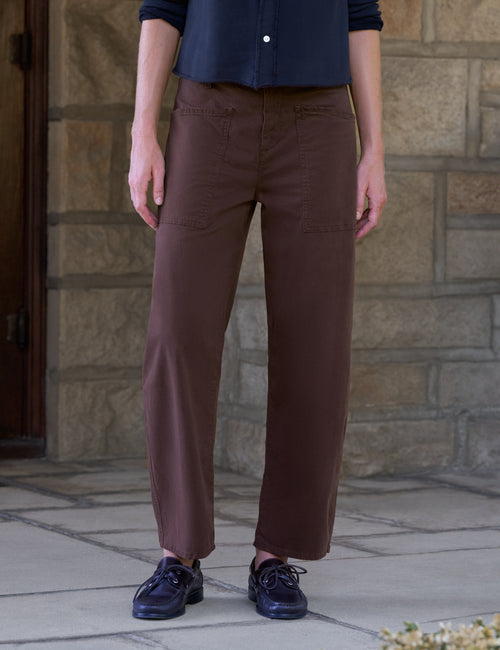 A person wearing brown barrel-leg trousers, a navy blue button-up shirt, and dark navy shoes, standing outdoors in front of a stone wall., front view