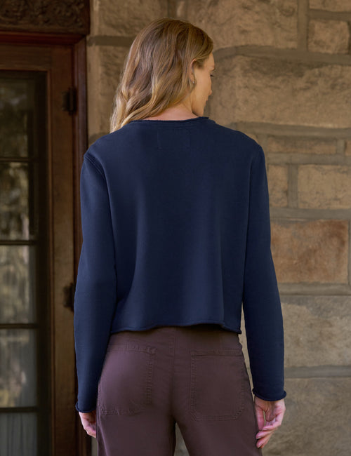 A woman shown from behind wearing a navy blue long-sleeve cropped sweater and brown pants, standing in front of a stone wall and a wooden door., alternate view
