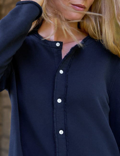 Close-up of a woman wearing a dark navy long-sleeve button-front top with raw, frayed trim along the neckline and front placket. The image is cropped from the lower face to the torso, with one hand near the hair., view 3